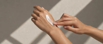Hands smoothing a small amount of white cream over the skin in soft sunlight